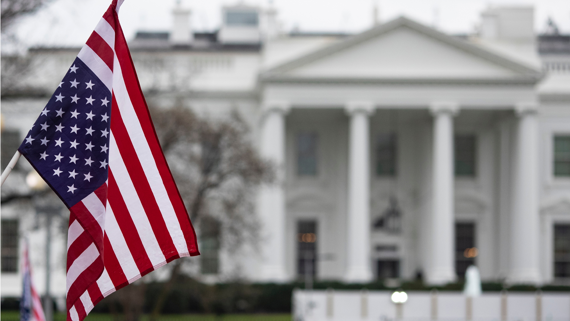 United States flag in the foreground with the White House blurred in the background, captured on a cloudy day.