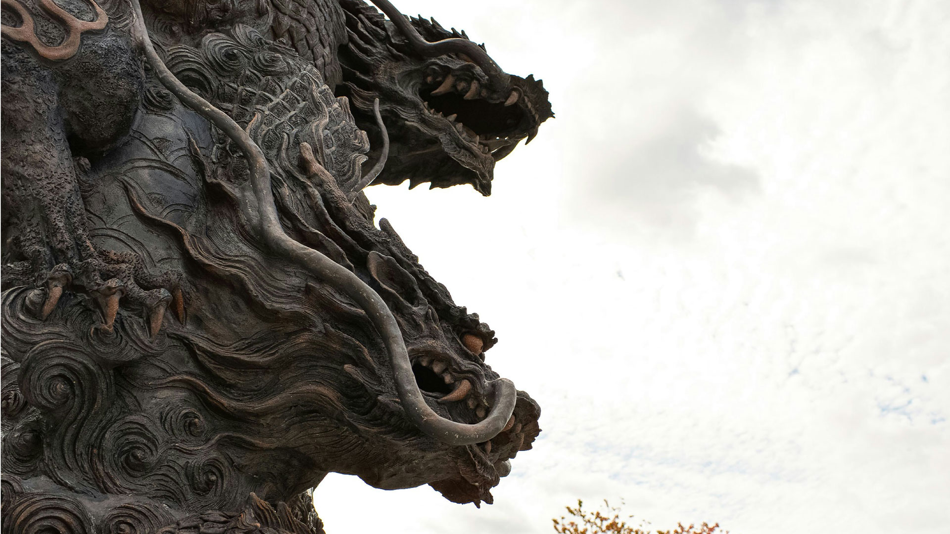 Close-up of a dramatic multi-headed dragon sculpture with detailed swirling textures, fierce expressions, and flowing mane, set against a cloudy sky.