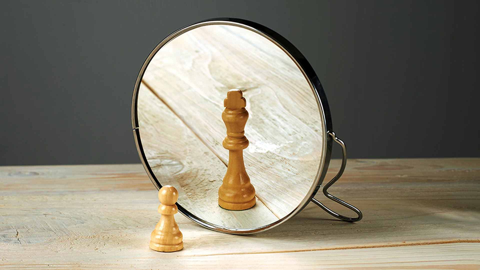 A small wooden chess pawn looking into a mirror and seeing the reflection of a king piece, symbolizing self-confidence, ambition, and inner potential.