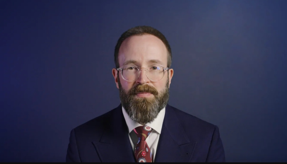 A bearded man with short dark hair and glasses, wearing a dark navy suit, white shirt, and a red patterned tie, poses in front of a plain dark blue background