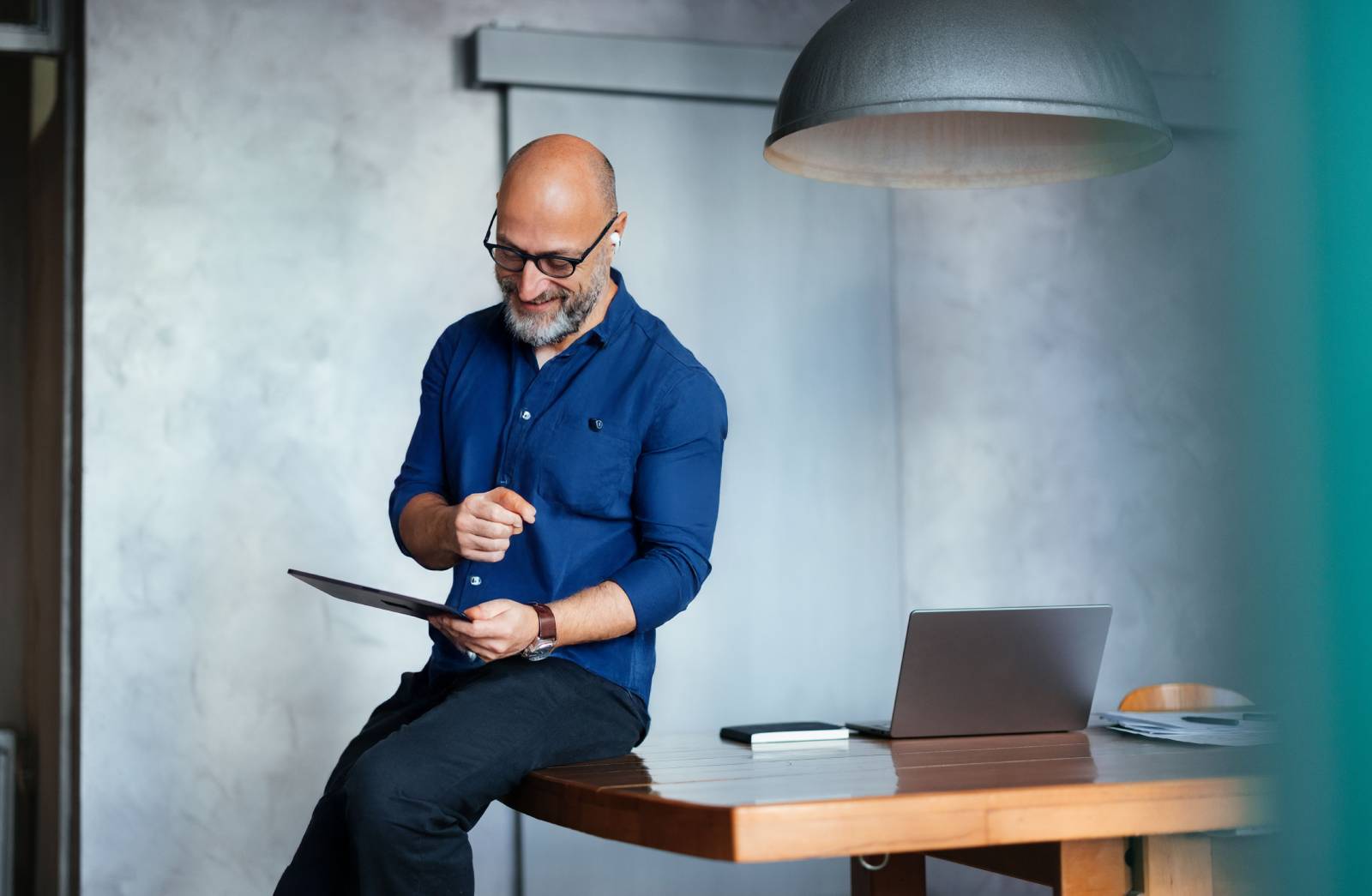 A man sits on the edge of a desk reading a tablet