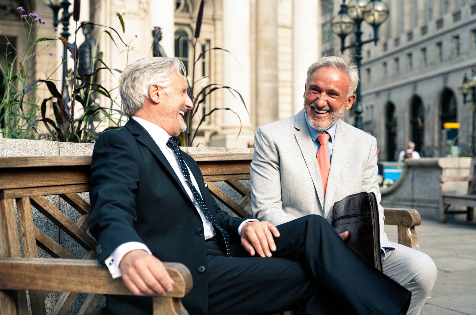 Two men in suits sit on a bench in the city having a conversation