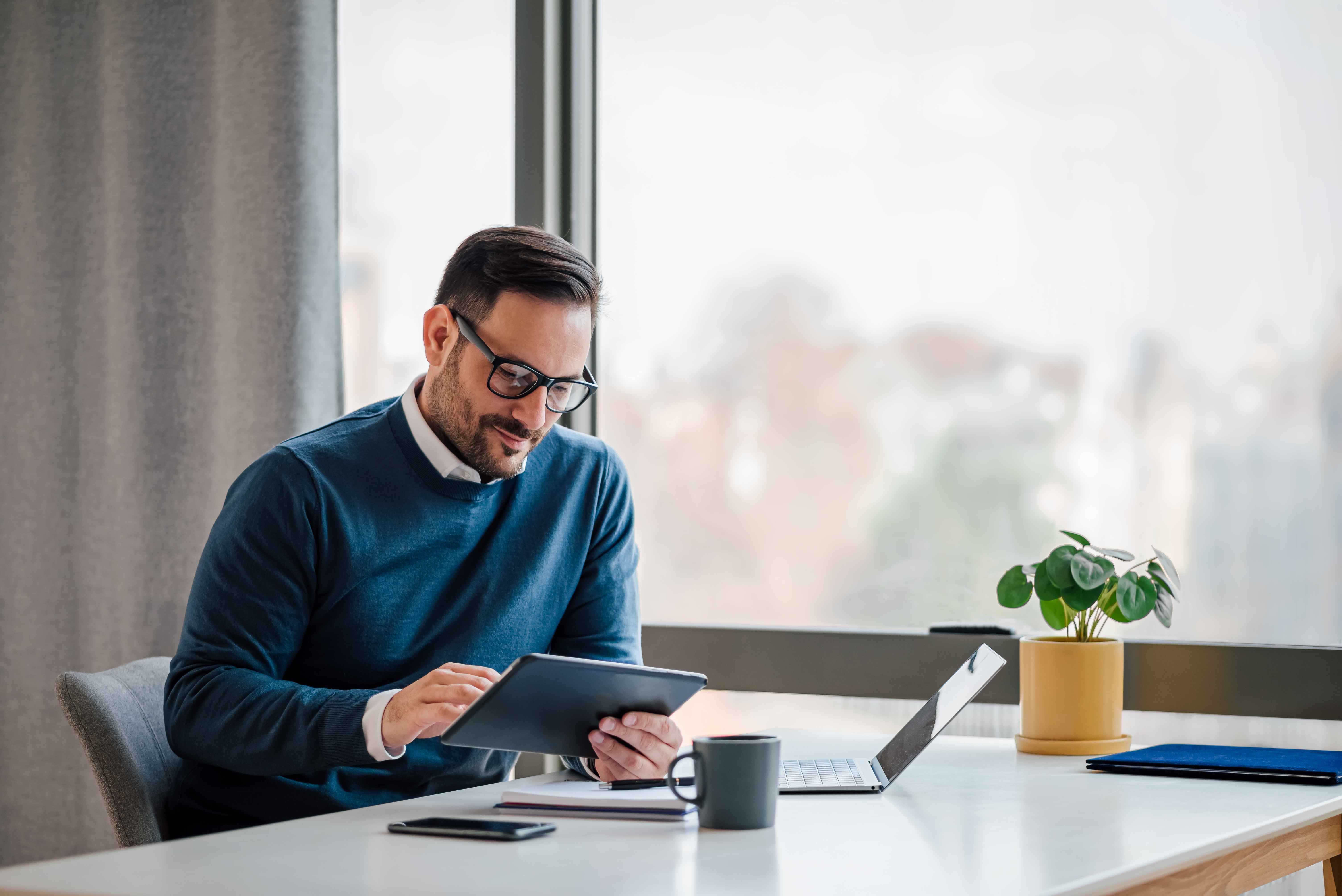A businessman working on a tablet with a laptop and coffee on his desk