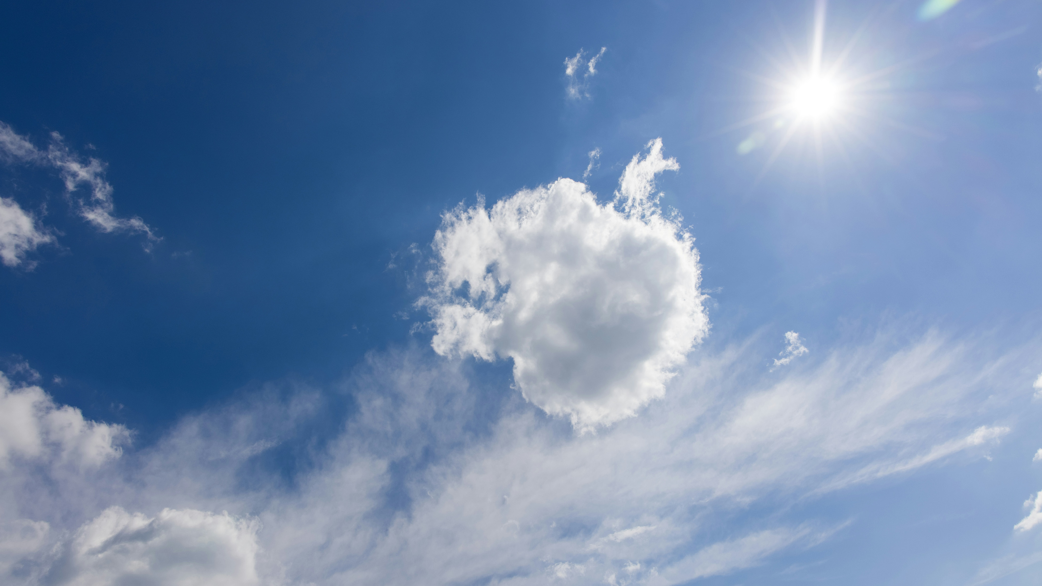 A bright blue sky with the sun shining in the top right corner and a single fluffy white cloud centered prominently, surrounded by wisps of thinner clouds.