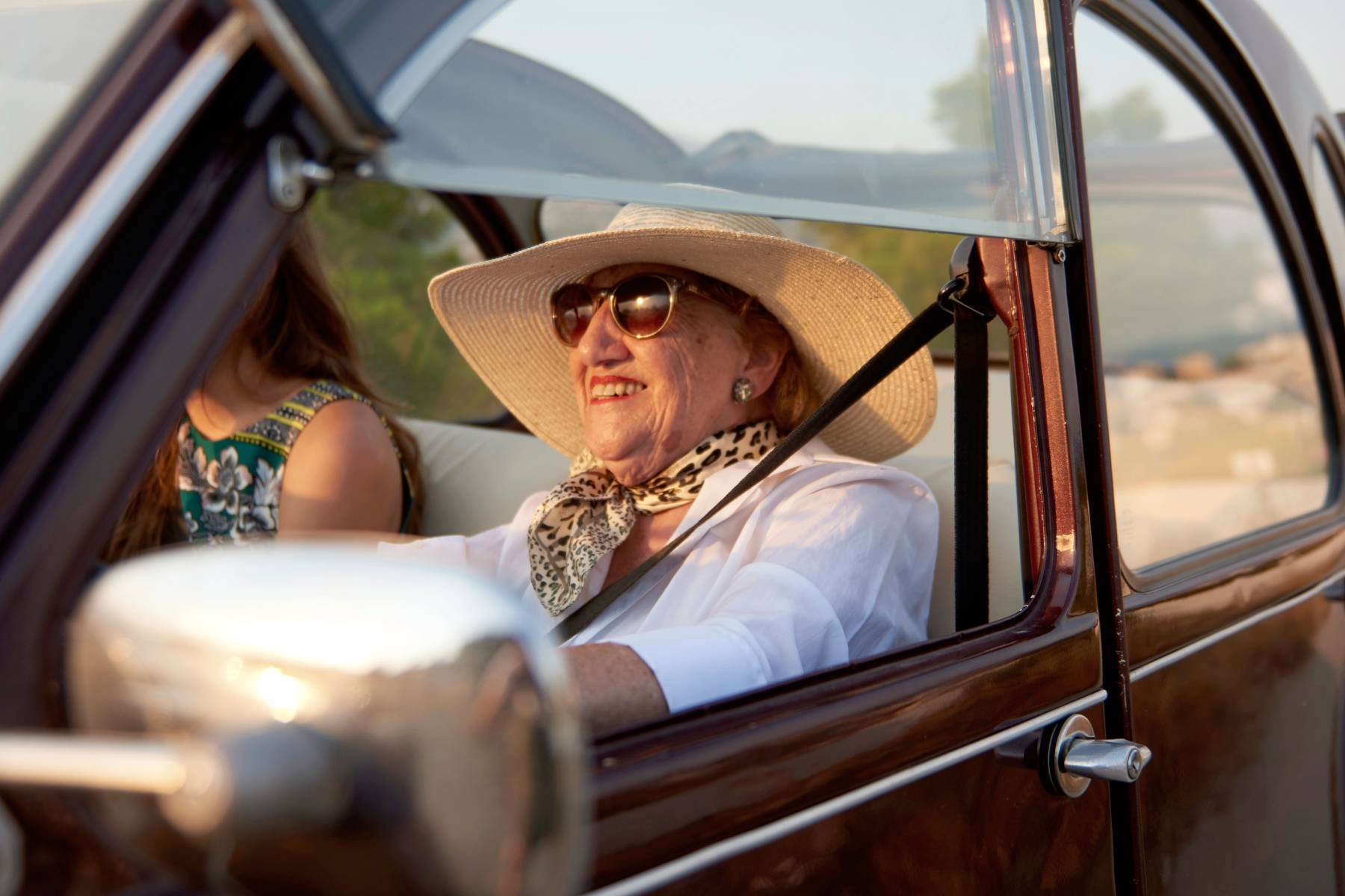 A lady wearing sunglasses and a sun hat smiles while driving in a vintage car
