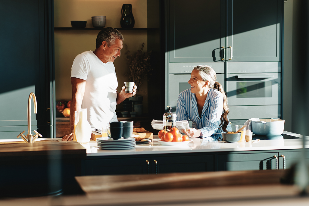 A couple talk together in kitchen while one sips coffee and the other reads the paper