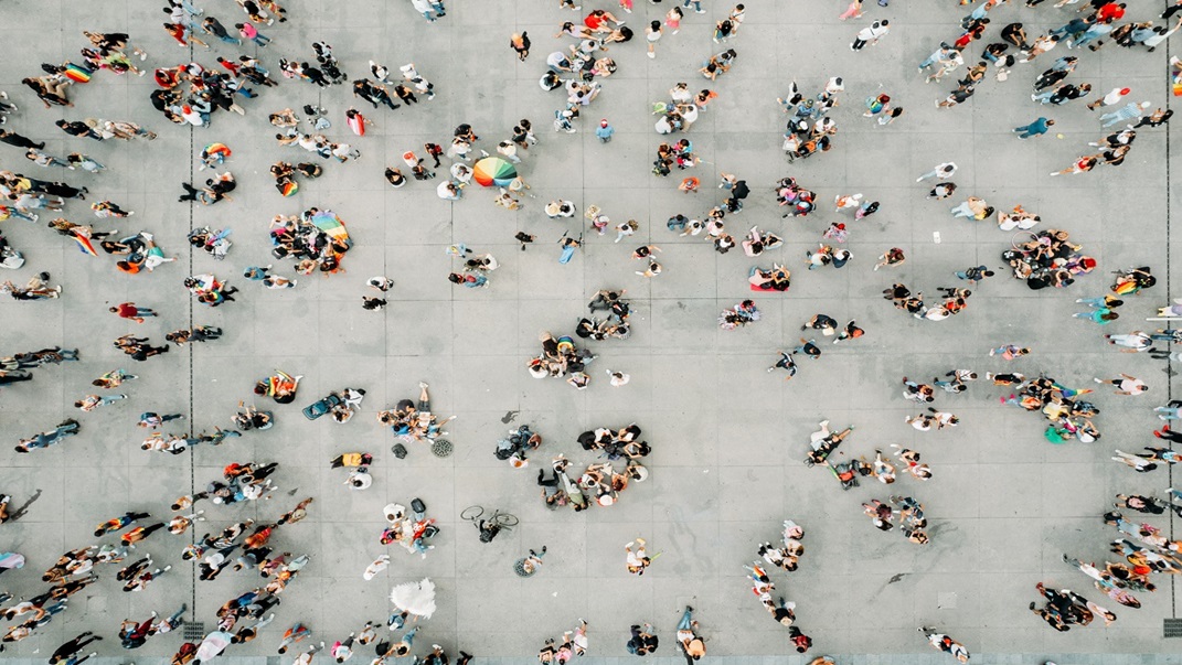 Aerial view of a large open plaza with many small groups of people scattered across the space, walking, standing, and interacting against a light-colored concrete surface
