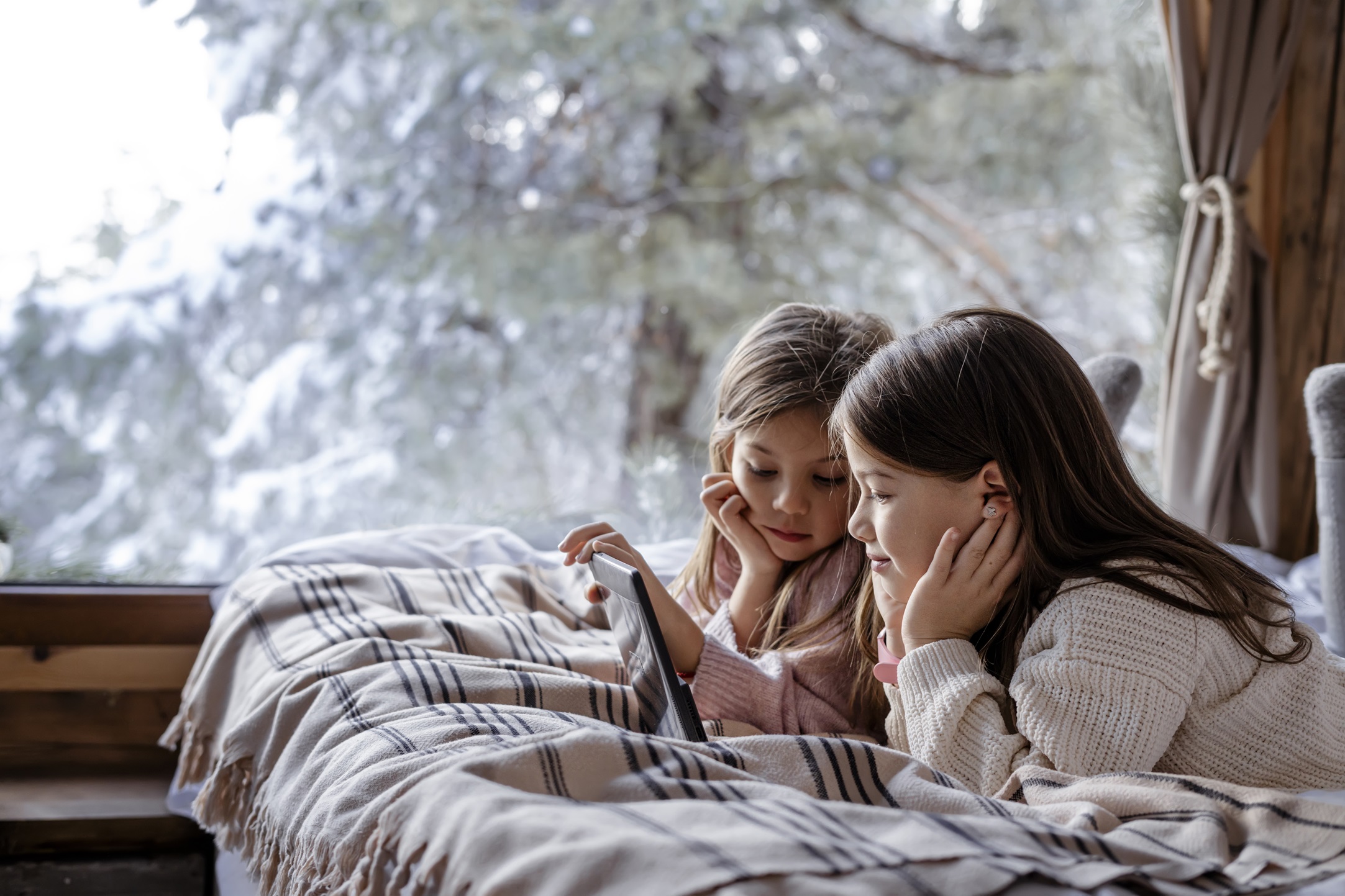 Two children lying under a plaid blanket indoors, watching a tablet near a large window with snowy trees outside