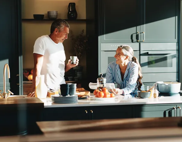 A couple talk together in kitchen while one sips coffee and the other reads the paper