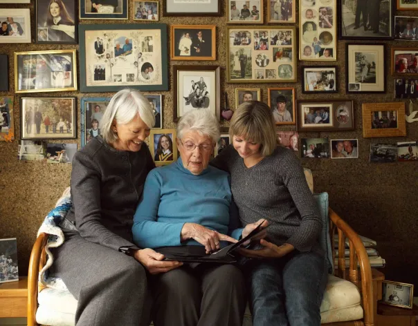 A mother and her grown up daughters sit together on a sofa and look at old family photos together