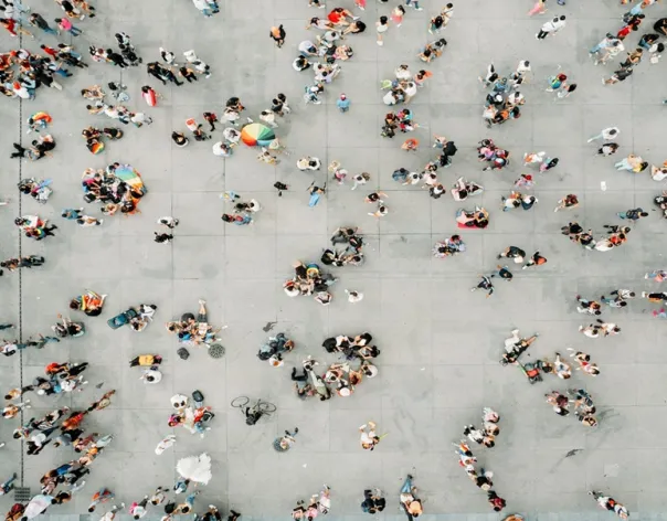 Aerial view of a large open plaza with many small groups of people scattered across the space, walking, standing, and interacting against a light-colored concrete surface