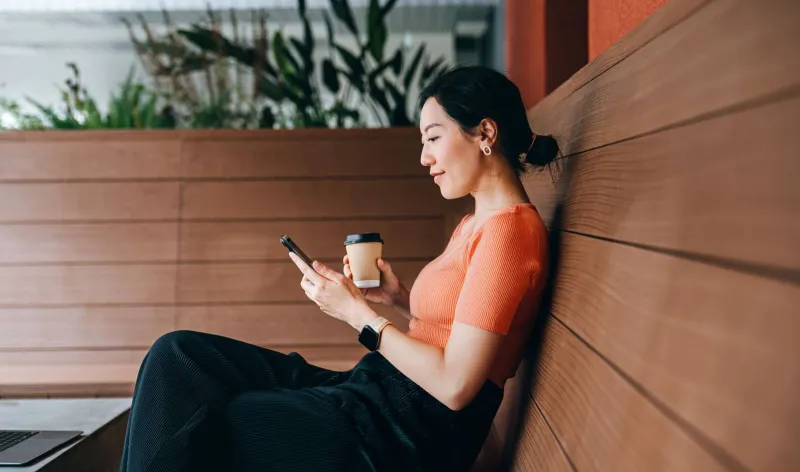A lady sits on a bench with mobile phone and coffee in hand