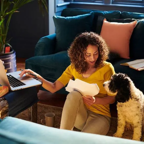 A lady sits on the floor with her dog while working on her laptop