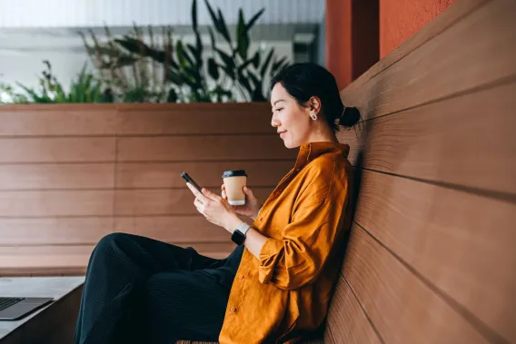 An accountant looking at her phone with a coffee in hand sitting on a bench