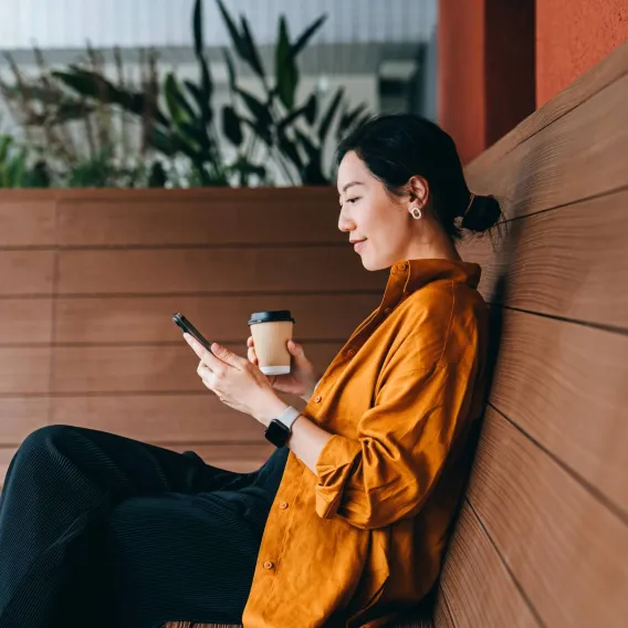 An accountant looking at her phone with a coffee in hand sitting on a bench