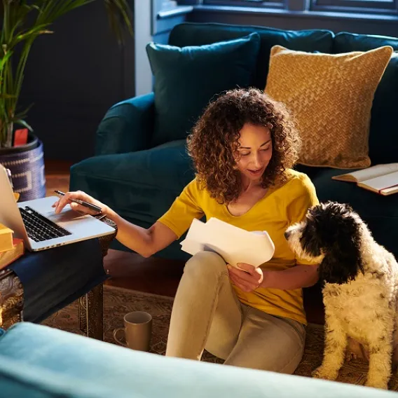 A lady sits on the floor of her house with her dog and a laptop