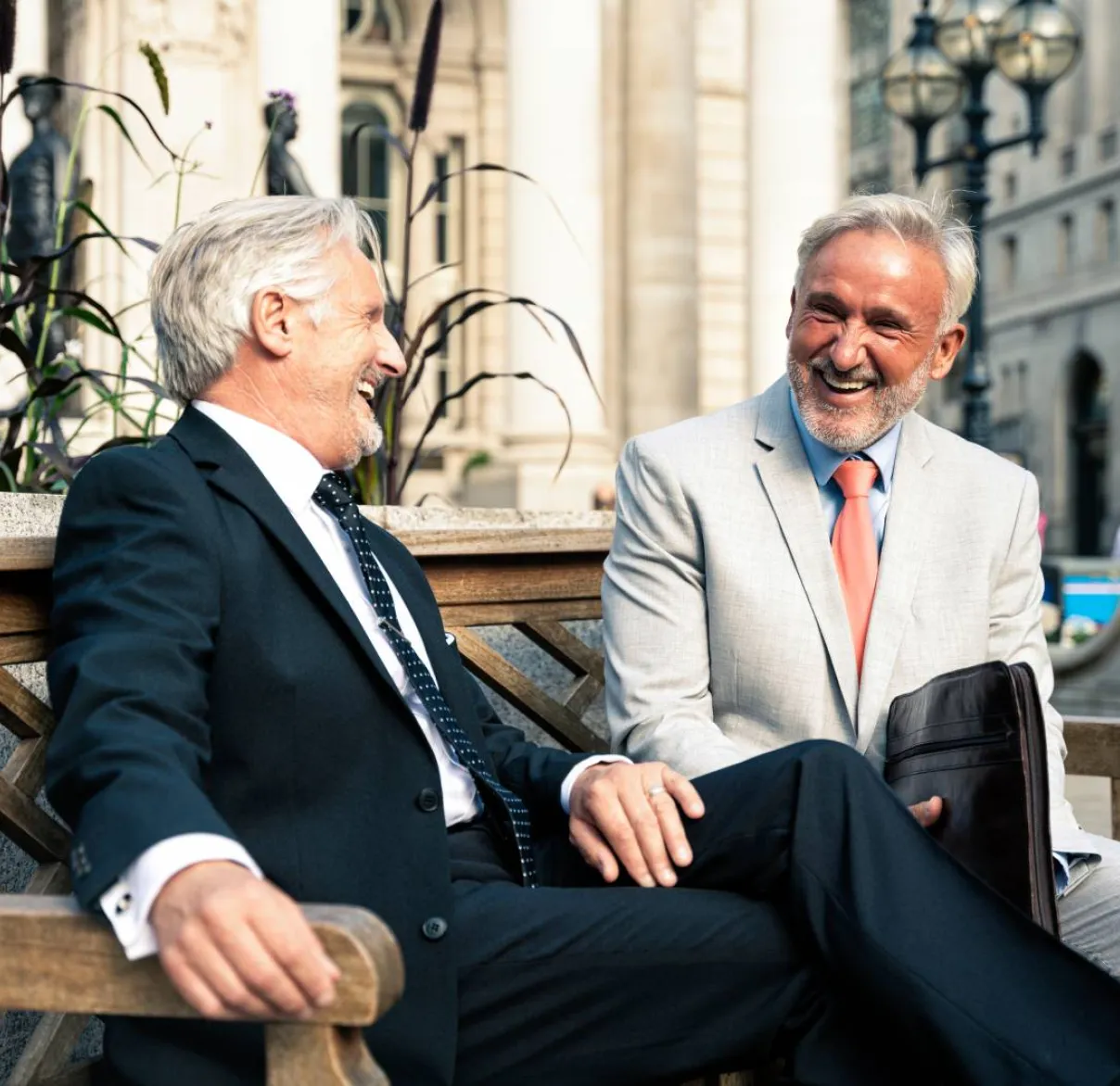 Two men in suits sit on a bench in the city having a conversation