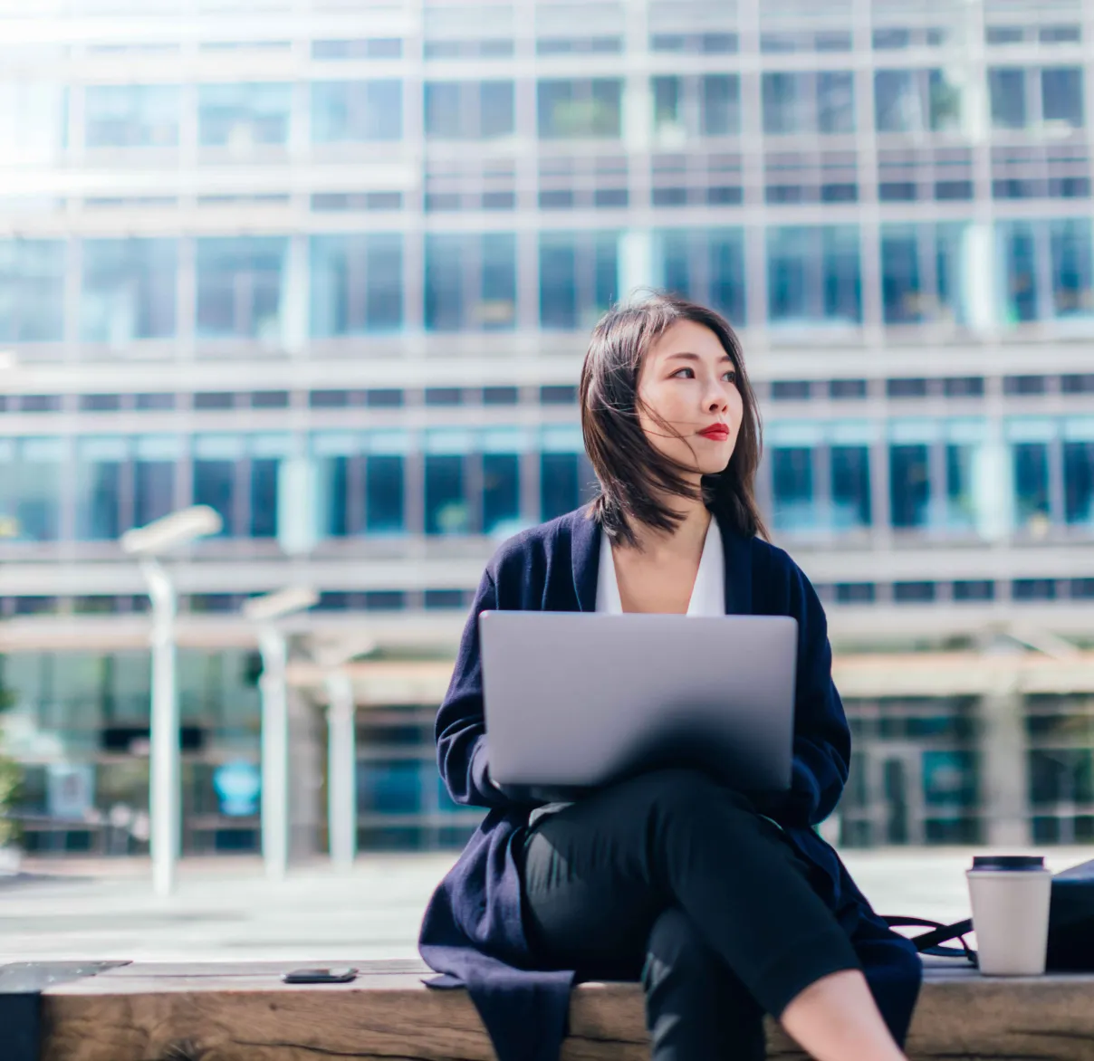 A woman sits on a bench outside a corporate office with a laptop on her leg