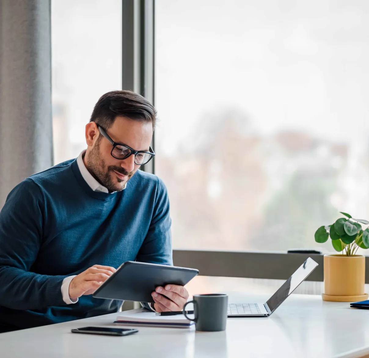An accountant sits at a desk with a laptop and a coffee cup and looks at something on his tablet