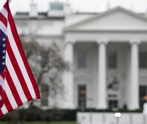 United States flag in the foreground with the White House blurred in the background, captured on a cloudy day.