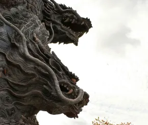 Close-up of a dramatic multi-headed dragon sculpture with detailed swirling textures, fierce expressions, and flowing mane, set against a cloudy sky.