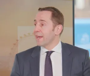 Man in a dark suit, white shirt, and dark tie speaking in a studio setting, with the London Eye faintly visible in the background and a potted plant on a shelf to the right.
