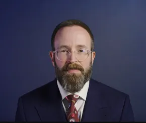 A bearded man with short dark hair and glasses, wearing a dark navy suit, white shirt, and a red patterned tie, poses in front of a plain dark blue background
