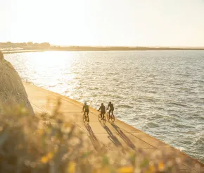 Three people cycle along the waterfront during sunrise