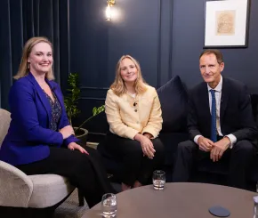 Three people seated in a studio-style room with dark blue walls, framed artwork and soft furnishings, with two people on a sofa and one in an armchair.
