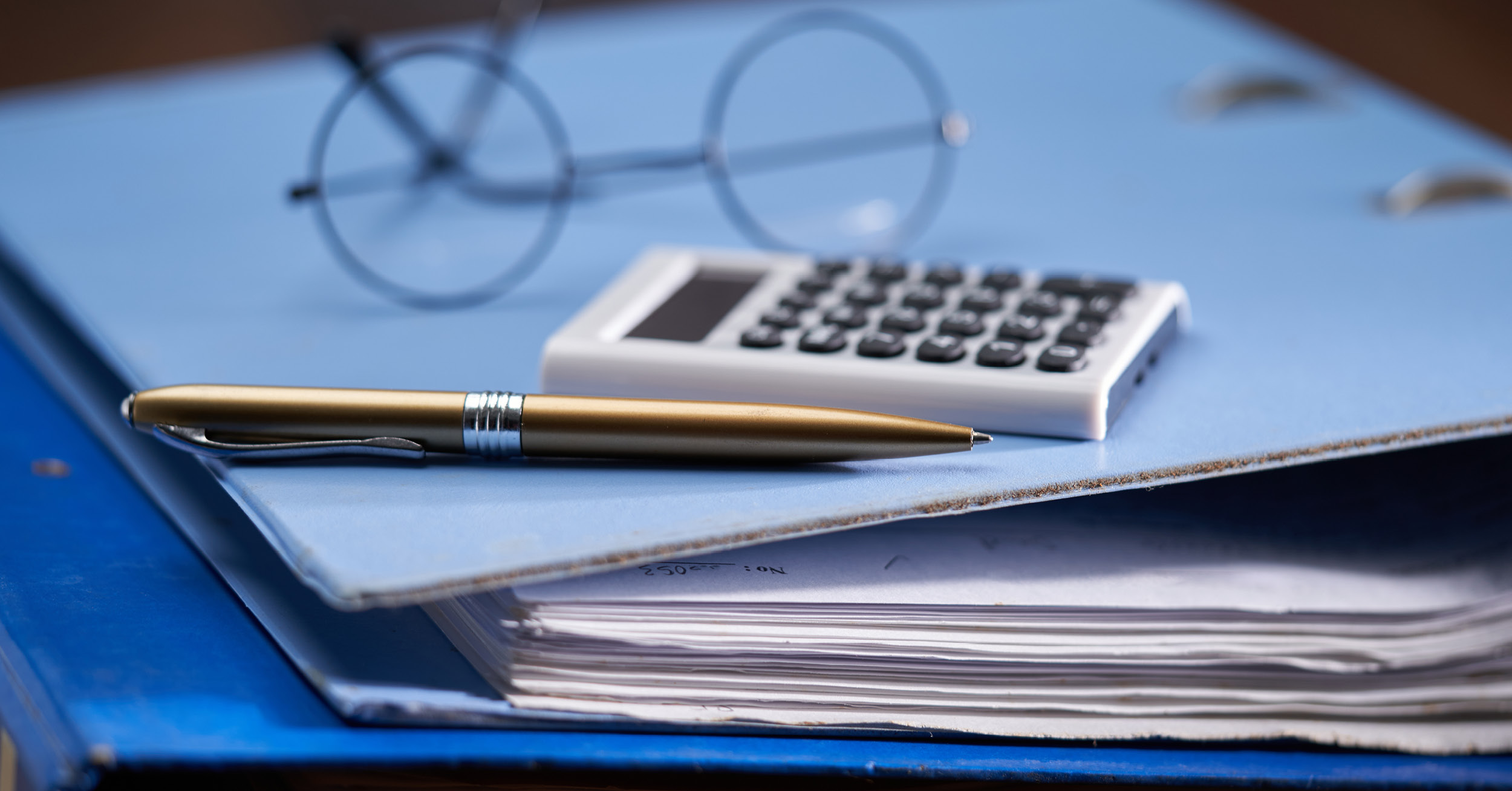 Black wire-framed spectacles, a white calculator and a gold pen lie on a light blue accounting ring-binder folder 